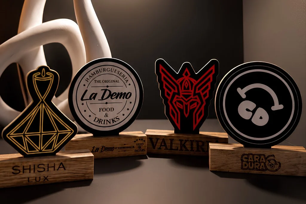 Cuatro soportes QR personalizados: bases de madera grabadas y placas bicapa en oro, blanco y rojo con formas a medida